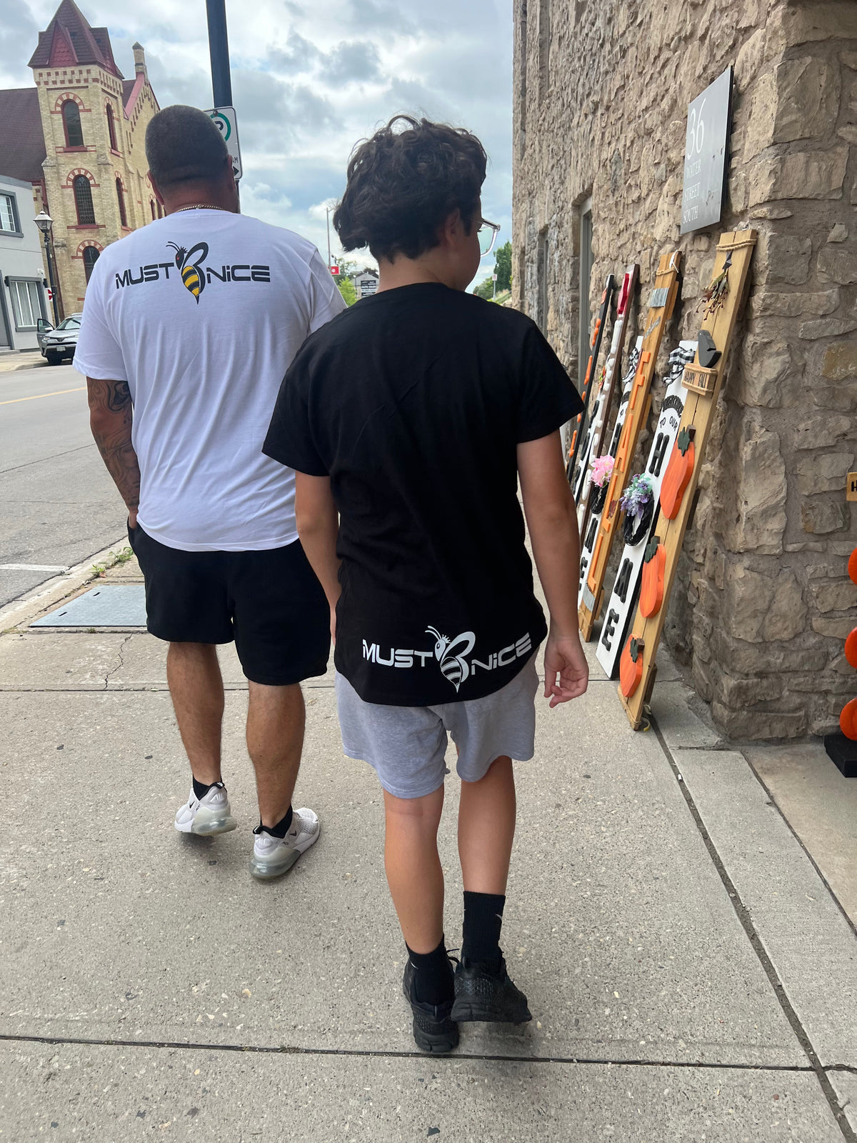 Father and son walking down a city sidewalk wearing Must Be Nice Essential Tees, passing a row of art panels or display boards along a stone building