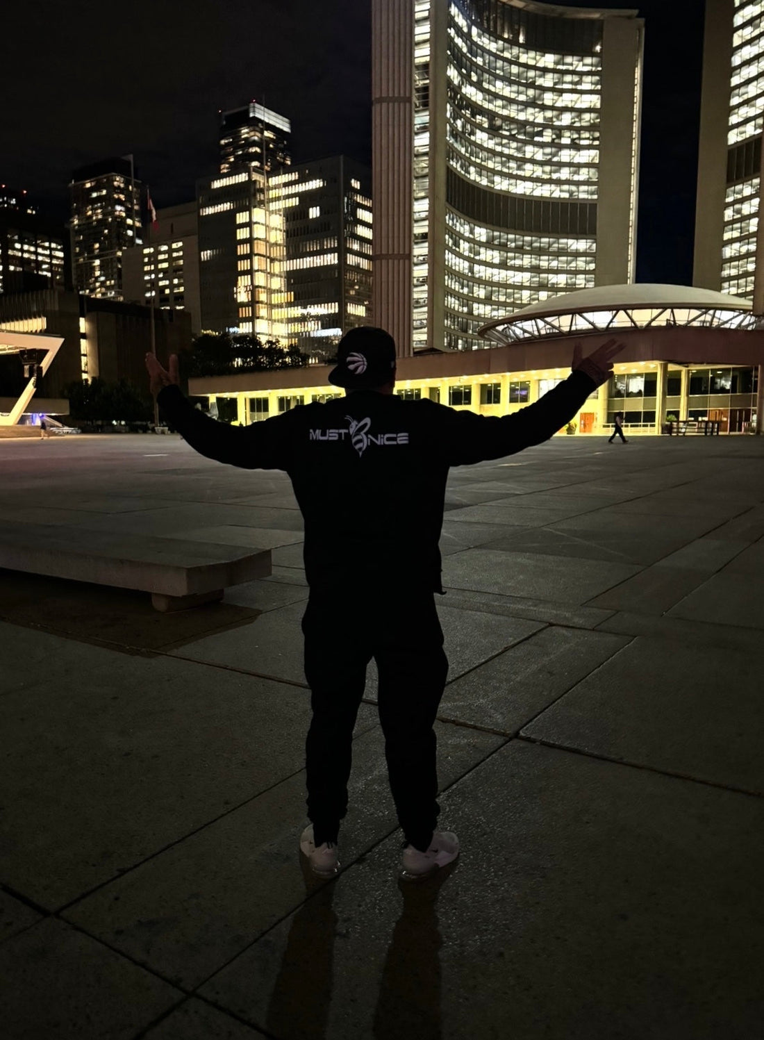 Man wearing a black Must Be Nice hoodie with arms raised at Toronto's Nathan Phillips Square at night, city hall towers lit up in the background