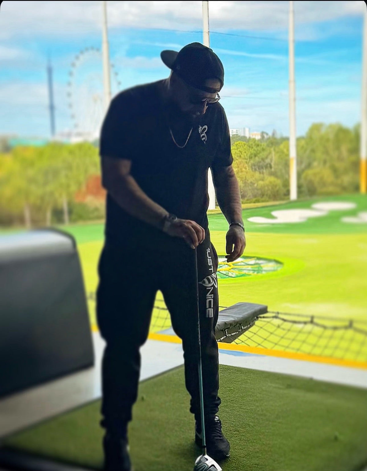 Man wearing black Must Be Nice bee logo joggers and t-shirt, driving a golf ball at an outdoor golf range