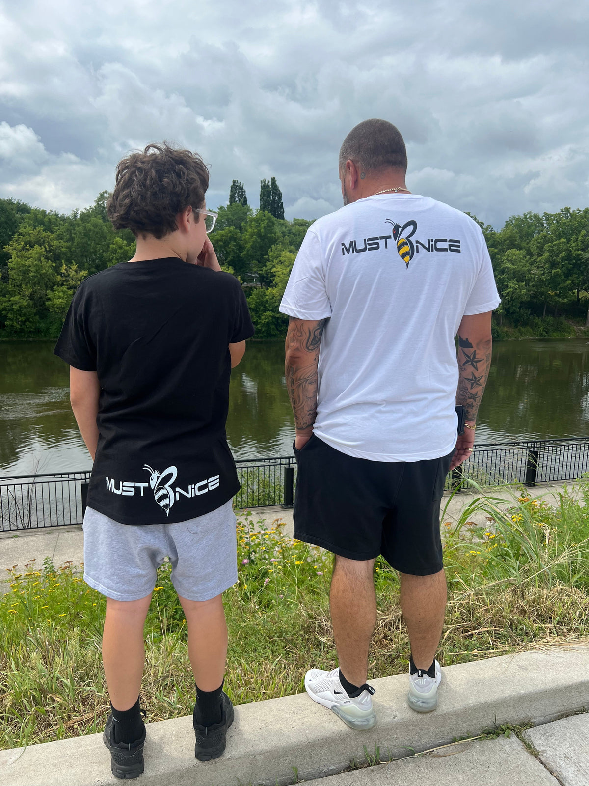Father and son wearing matching Must Be Nice t-shirts with printed bee logo, standing by a river